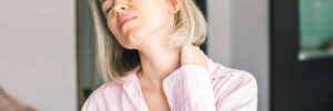 A person with short blonde hair wearing a pink shirt appears to be experiencing neck pain, holding their neck and grimacing while standing indoors.