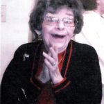 Elderly woman with short curly hair and glasses, wearing a black jacket with red accents, clapping her hands and smiling widely indoors. .