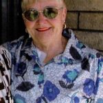 An older woman wearing sunglasses, a patterned blouse with blue flowers, and gold earrings smiles while standing against a brick wall.